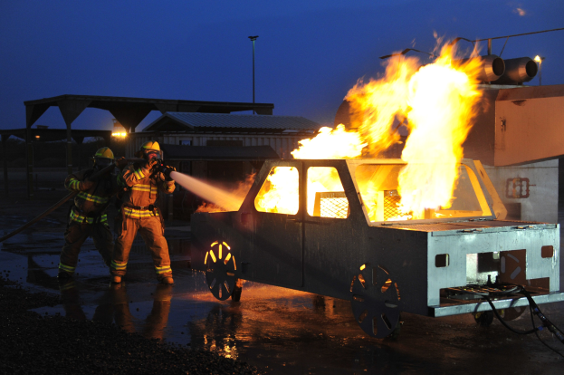 Feuerwehrleute mit Helmen stehen vor einem lodernden Truck, halten Schläuche, mit Schuppen, Pfählen, Lampen, Bäumen und einem klaren blauen Himmel im Hintergrund.