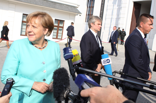 Bundeskanzlerin Angela Merkel spricht vor dem Kanzleramt in Berlin mit Reportern, umgeben von Menschen mit Mikrofonen und Gebäuden im Hintergrund.