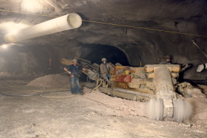 Gruppe von Männern in Helmen, die in einem dunklen Stollennetz mit Maschinen und Rohren arbeiten.