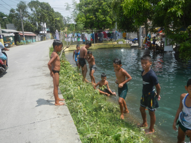 Kinder spielen am Straßenrand in flachem Wasser, einige stehen, während andere auf dem Gras sitzen, mit einer Person auf einem Motorrad links und Häusern, Bäumen und Strommasten im Hintergrund unter einem klaren blauen Himmel.
