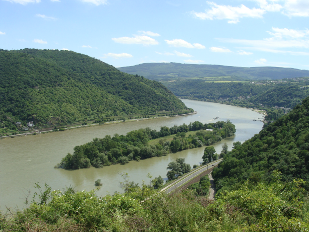 Panoramablick auf den sich durch eine grüne Landschaft schlängelnden Rhein mit einer Straße daneben unter einem teilweise bewölkten Himmel.