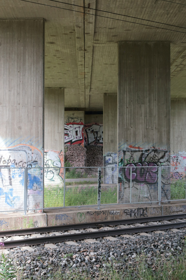 Eisenbahnschiene mit Graffiti an einer Betonwand, umgeben von Steinen, Gras, einem Zaun, Pfeilern, Oberleitungen und einem Dach.