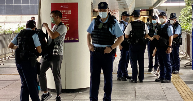 Gruppe von Polizisten in Uniform mit Mützen und Gesichtsmasken vor einem Gebäude stehend, einige halten Kameras, mit einem Banner an einer Säule, Geländern, Pflanzen, Bäumen und Deckenlampen im Hintergrund.