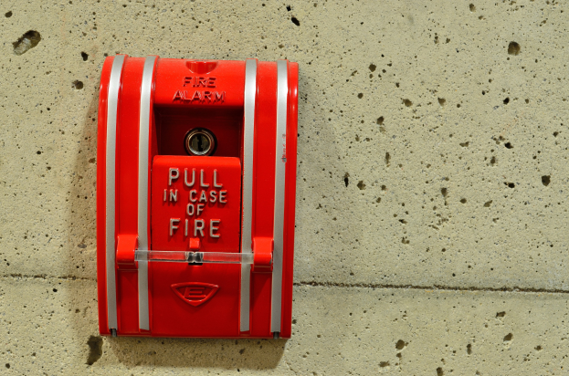 Roter zylindrischer Feueralarmkasten mit glänzender Oberfläche, einem oberen Griff und einem mittigen Schlüsselloch, montiert an einer Gebäudeseite mit der Aufschrift "Feueralarm" in fetter schwarzer Schrift.