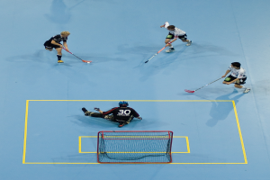 Drei Personen in Sportkleidung spielen Hockey, wobei eine Person rechts mit einem Hockeystock den Ball schlägt und eine Person hinter einem Tor auf dem Boden sitzt.