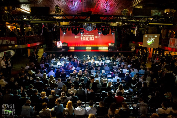 Eine große Menschenmenge sitzt auf Stühlen vor einer Bühne mit Bildschirmen und Bannern, die von Scheinwerfern beleuchtet wird und von Geländern umgeben ist, und wartet gespannt auf den Beginn eines Online-Marketing-Events für Rockstars, mit einem Wasserzeichen in der linken unteren Ecke.