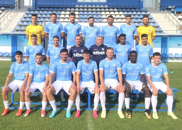 Gruppe von Männern auf einem Fußballfeld, einige sitzen auf Bänken und andere stehen auf dem Rasen, mit Tribünen und Strukturen im Hintergrund.