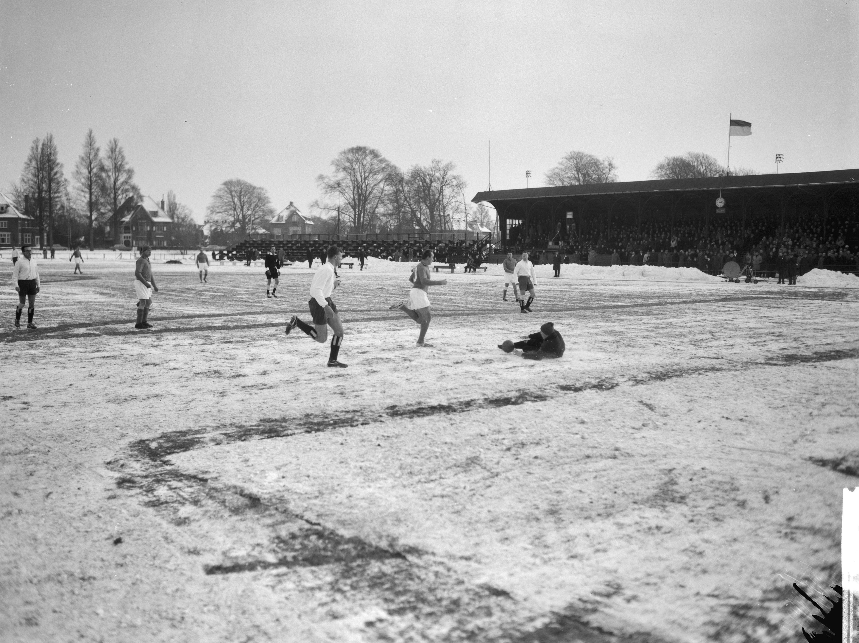 Schwarzes Foto von Menschen, die auf einem schneebedeckten Fußballfeld spielen, mit Häusern, Bäumen, Pfählen, Fahnen und einer Hütte im Hintergrund.