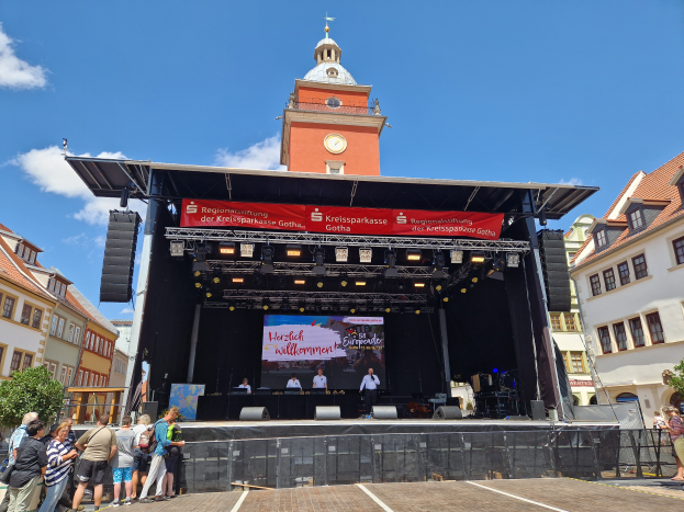 Eine Gruppe von Menschen auf einer Bühne vor einem Uhrenturm, umgeben von Gebäuden, Bäumen und einem bewölkten Himmel, mit einem Bildschirm auf der Bühne, beim Kreissparkasse Gotha Musikfestival.