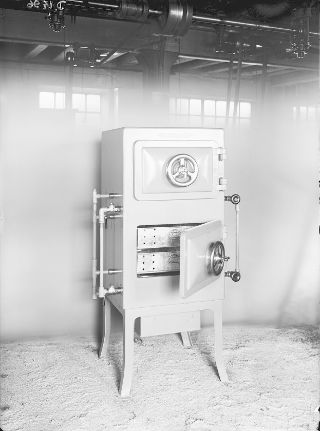 Schwarzes und weißes Foto eines altmodischen Safes in einem Raum mit einer Wand, Fenstern und an der Wand entlanglaufenden Rohren.