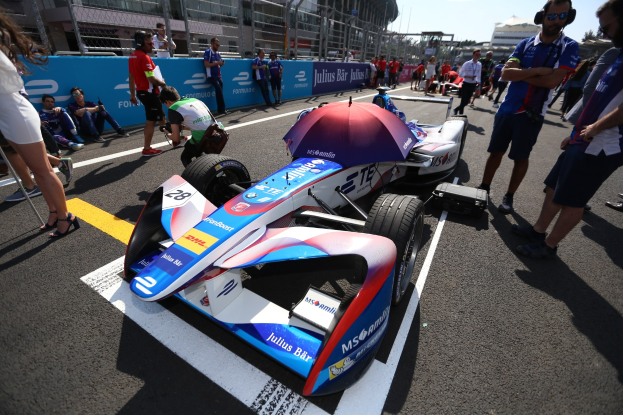Formula E-Rennwagen mit weißer, blauer, roter und schwarzer Lackierung, der auf einer Strecke geparkt ist, umgeben von Menschen, mit einem Schirm im Vordergrund und Gebäuden, Bäumen und einem klaren Himmel im Hintergrund.
