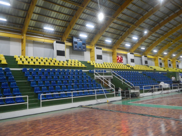 Großer Hallensportplatz mit Holzboden, blauen und gelben Sitzplätzen, Geländern, Treppen zu den Sitzplätzen, Wandbildschirme, Deckenleuchten und einigen anwesenden Personen.