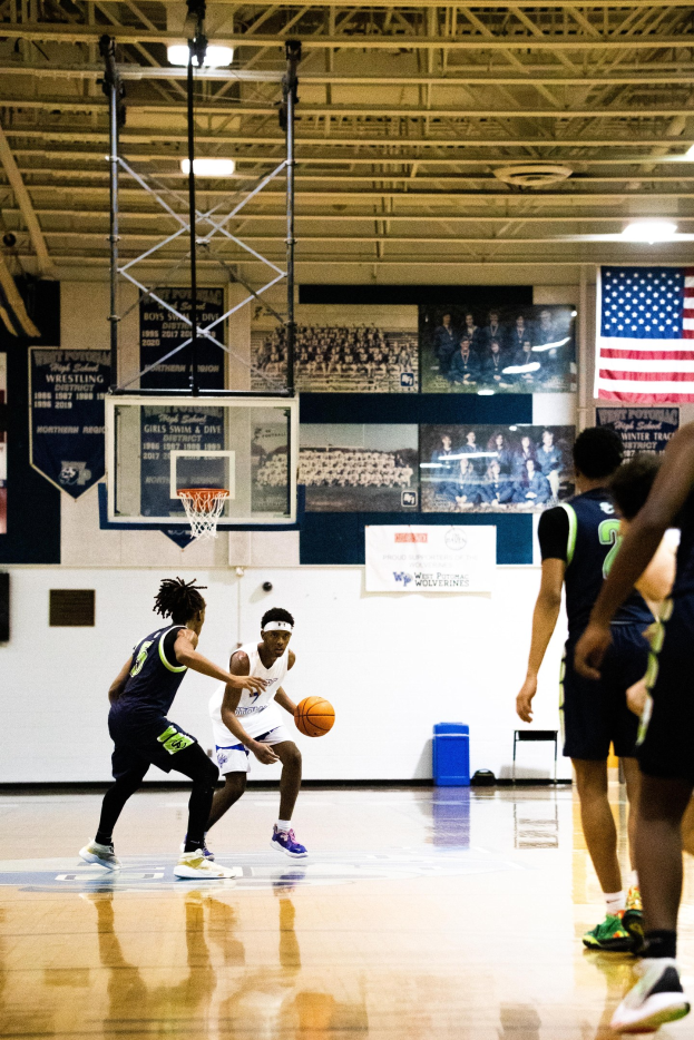 Junge Männer spielen Basketball in einer Halle mit einer Wand, Deckenlampen und einer Flagge auf der rechten Seite.