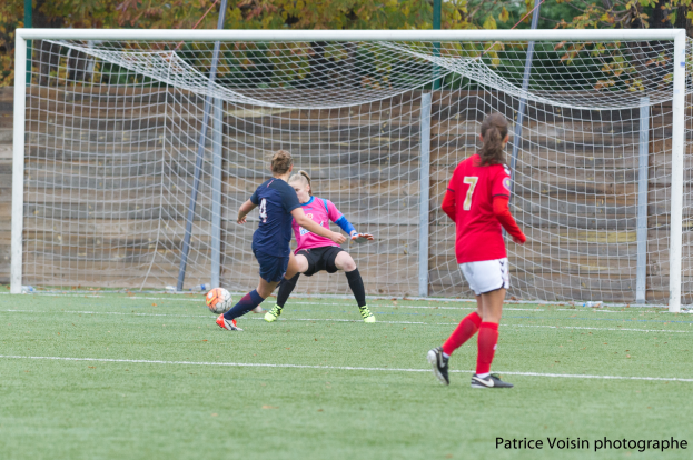 Eine Gruppe von Frauen, die Fußball auf einem Rasenfeld umgeben von Bäumen spielen, mit einem Torpfosten im Hintergrund und Text am unteren Rand des Bildes.