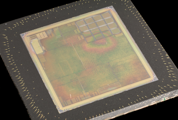 Nahaufnahme eines rechteckigen Halbleiterchips mit metallischem Glanz, umgeben von einem schmalen Metallrahmen und mit kleinen Löchern an seinen leicht erhöhten Kanten auf schwarzem Hintergrund.