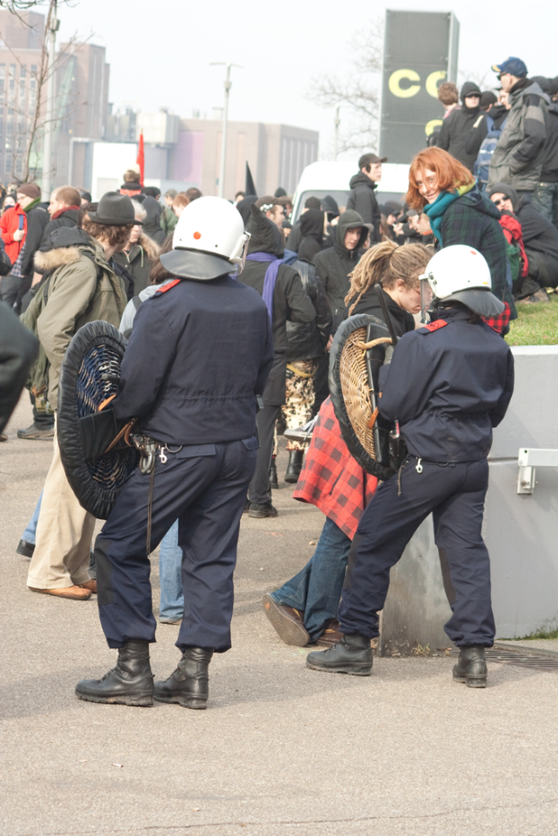 Eine Gruppe von Menschen, die auf einer Straße mit zwei Personen in der Mitte gehen, die wie Polizisten aussehen, im Hintergrund Gebäude und unten Boden.