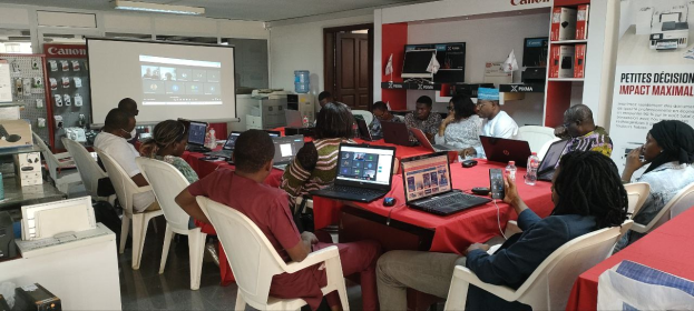 Gruppe von Menschen an einem Tisch mit Laptops, Flaschen und anderen Gegenständen während eines digitalen Marketing-Workshops, mit einem Banner, einem Display-Bildschirm, einer Tür und Deckenleuchten im Raum.