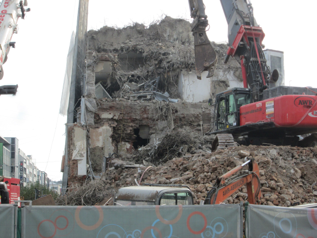 Ein Kran im Vordergrund zerstört ein Gebäude, umgeben von anderen Kränen, Fahrzeugen, Felsen und einem Banner mit Text, mit Gebäuden, Bäumen und Himmel im Hintergrund.