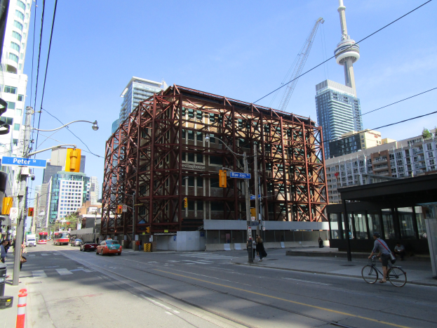 Eine belebte Stadtstraße mit einem großen Gebäude im Hintergrund, Fahrzeugen auf der Straße, einem Radfahrer, Fußgängern auf dem Gehweg, Verkehrsampeln, Pfosten mit Schildern, einem Kran und dem Himmel.