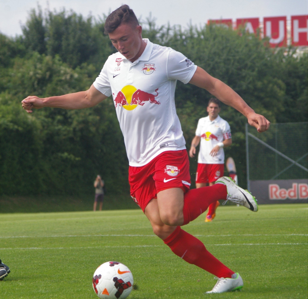 Ein Profifußballer tritt einen Fußball auf einem grünen Feld mit "RB Leipzig" im Hintergrund unter einem klaren blauen Himmel.