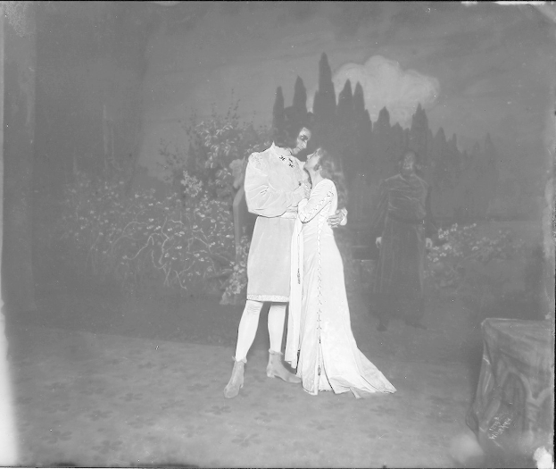 Schwarzes und weißes Foto eines Mannes und einer Frau, die sich auf einer Bühne umarmen und küssen, mit ein paar Menschen, Pflanzen und Bäumen im Hintergrund.