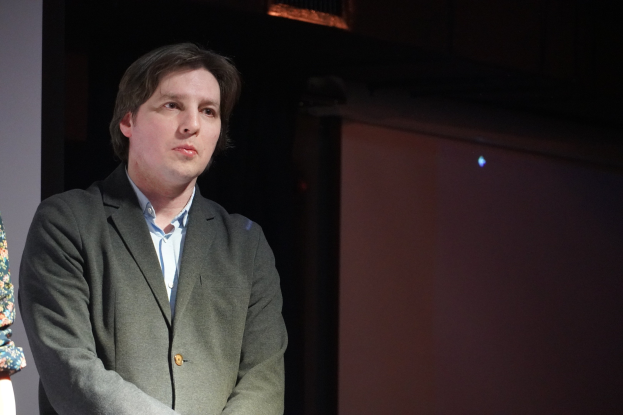 Ein Mann im Anzug und eine Frau stehen nebeneinander auf einer Bühne, beide mit ernsten Gesichtern, als würden sie über etwas Wichtiges diskutieren.