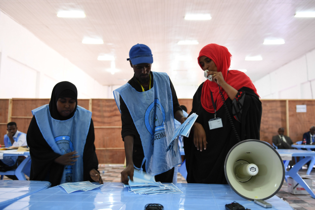 Eine Gruppe von Menschen um einen Tisch mit Megafon und Papieren, mit anderen im Hintergrund unter einer beleuchteten Decke, in einer Wahllokal.