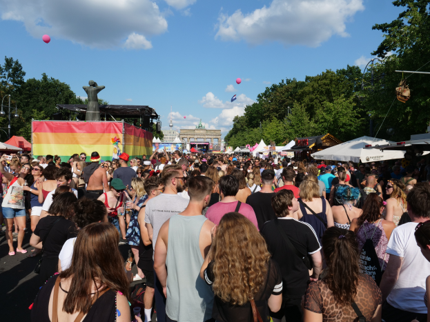 Eine große Menschenmenge, die eine Straße mit Zelten, Bäumen, Pfählen, Lichtern und einer Statue gesäumt, im Hintergrund Gebäude und ein Himmel mit Wolken und Ballons, während des Berliner Pride-Festivals.