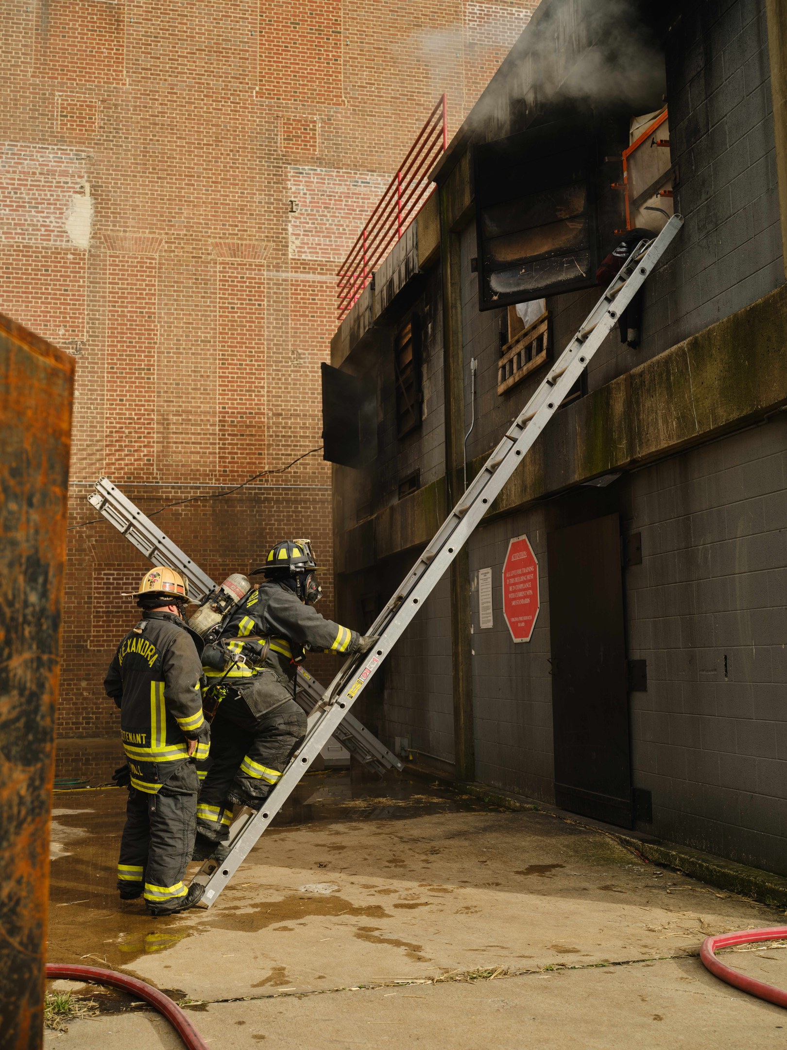 Feuerwehrleute mit Helmen klettern eine Leiter zu einem rauchgefüllten Gebäude hoch, während in der Nähe ein metallisches Objekt und Bodenschläuche sichtbar sind.