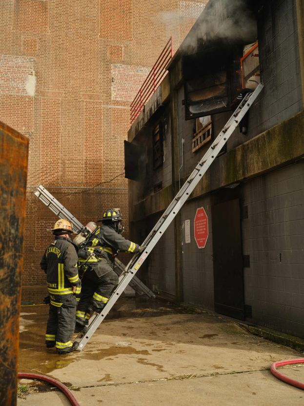Feuerwehrleute mit Helmen klettern eine Leiter zu einem rauchgefüllten Gebäude hoch, während in der Nähe ein metallisches Objekt und Bodenschläuche sichtbar sind.