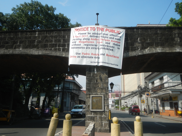 Eine Brücke mit einem "Hinweis an die Öffentlichkeit"-Schild, Fahrzeuge auf der Straße darunter und Gebäude, Bäume, Masten, Drähte und einen klaren blauen Himmel im Hintergrund.