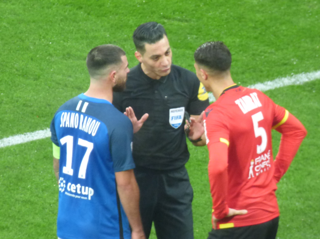 Schiedsrichter spricht mit zwei Männern auf einem Fußballfeld; einer trägt ein schwarzes Hemd und Hosen, der andere ein blau-rotes T-Shirt mit Text, beide tragen eine Brille.
