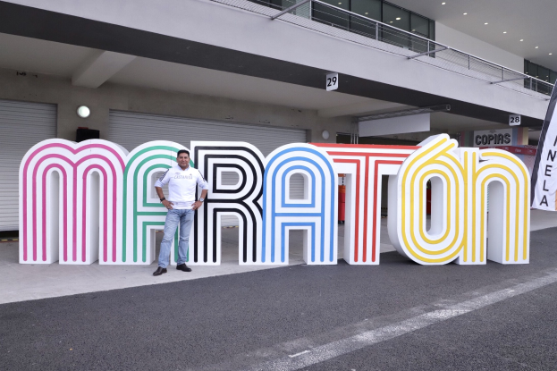 Ein Mann steht vor einem großen "Marathon"-Schild mit einem Banner auf der rechten Seite, mit einem Gebäude mit Lichtern, Geländern und Läden im Hintergrund.