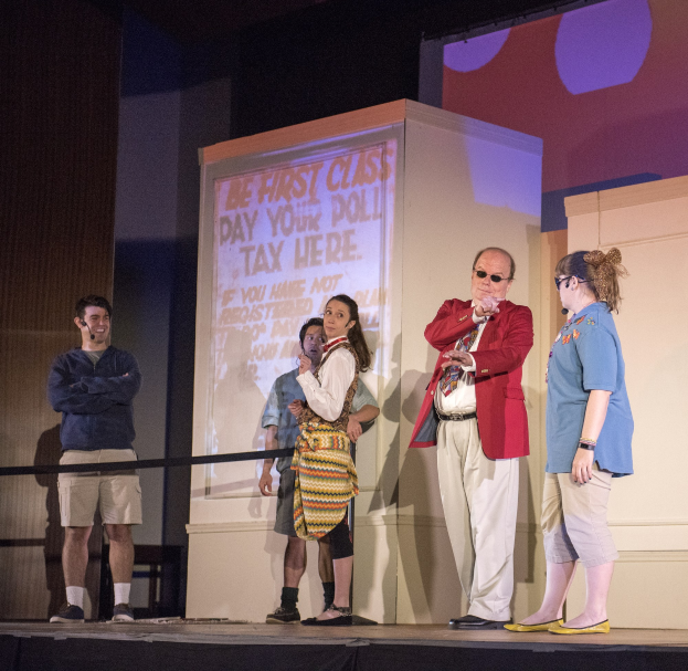 Gruppe von Menschen auf einer Bühne mit einem Mann, der ein Mikrofon hält, einem Bildschirm mit der Aufschrift "The First Class Day Your Poll Tax Here", einem Zaun, einer Wand und Deckenlampen.