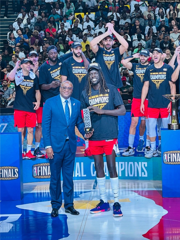 Gruppe von Männern mit Trophäen auf einem Basketballfeld mit Zuschauern im Hintergrund und zwei Trophäen auf Podesten.