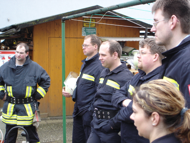 Eine Gruppe von Männern in blauen Feuerbekämpfungsuniformen steht nach links gewandt, mit einem kleinen hölzernen Schuppen und Rohrrahmen im Hintergrund.