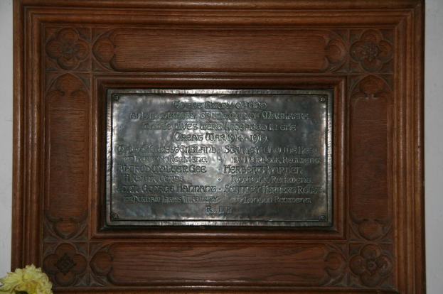 Eine Gedenktafel mit Text ist an einer braunen Wand oder einer hölzernen Tür angebracht, mit einem gelben Blumenstrauß in der unteren linken Ecke.