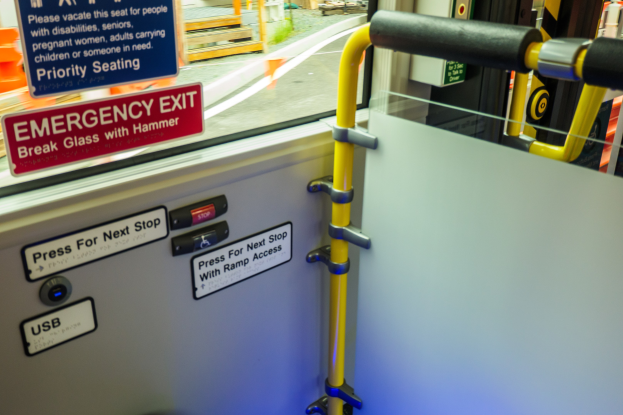 Notausgangsschild an der Seite eines Zuges mit Tür, Griff und Glasfenster, das Straße, Pfosten und andere Objekte draußen zeigt.