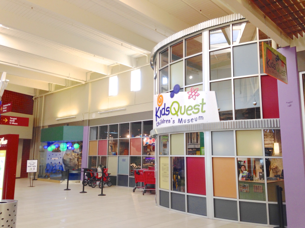 Interior of a shopping mall with a "Kids Quest Children's Museum" sign, featuring trolleys, ribboned poles, text boards, ceiling lights, and scattered objects.