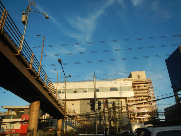 Stadtstraße mit parkenden Autos, eine Brücke mit Geländern, Laternen, Strommasten mit Drähten, Ampeln, Gebäude mit Fenstern und ein bewölkter Himmel im Hintergrund.