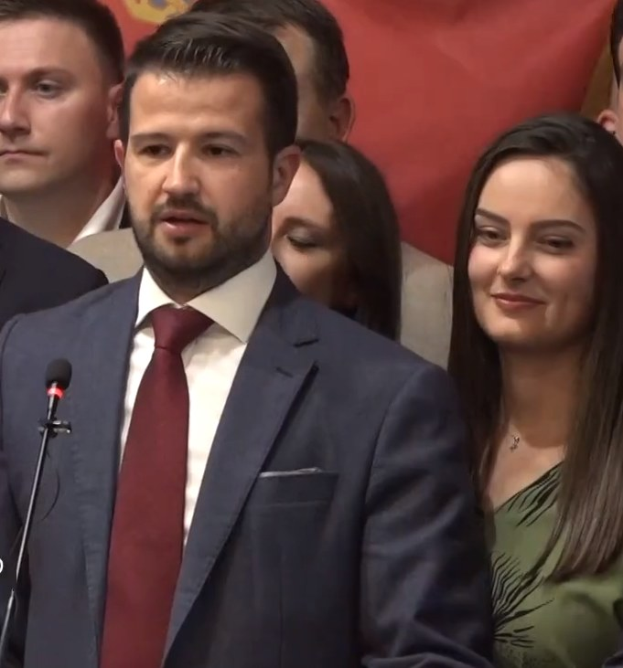 Ein Mann im Anzug spricht in ein Mikrofon auf einem Podium, mit einem Banner und Publikum im Hintergrund, wahrscheinlich zu einem Regierungsverbot von sozialen Medien.