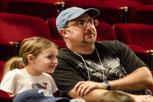 Ein Mann mit Brille, Uhr und Mütze sitzt neben einem strahlenden kleinen Mädchen in einem Kino, im Hintergrund sind andere Menschen und Stühle zu sehen.