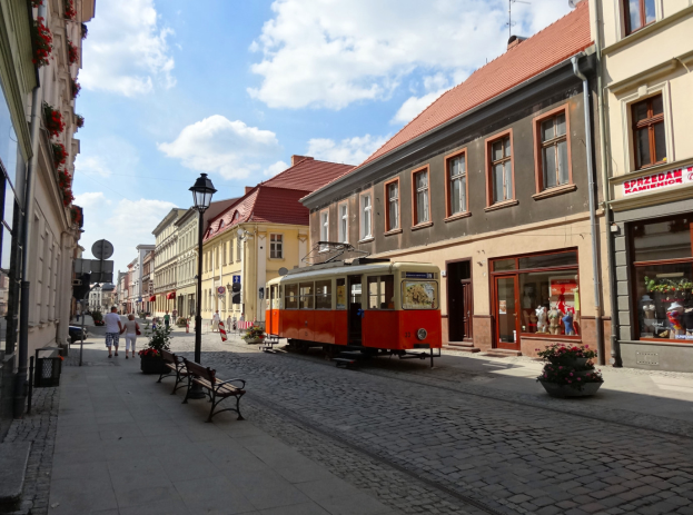Eine rote und gelbe Tram fährt durch die Kopfsteinpflasterstraßen der Altstadt von Tallinn, flankiert von Gebäuden mit Fenstern, Bänken und Laternen, mit Menschen auf den Bürgersteigen und einem bewölkten Himmel im Hintergrund.