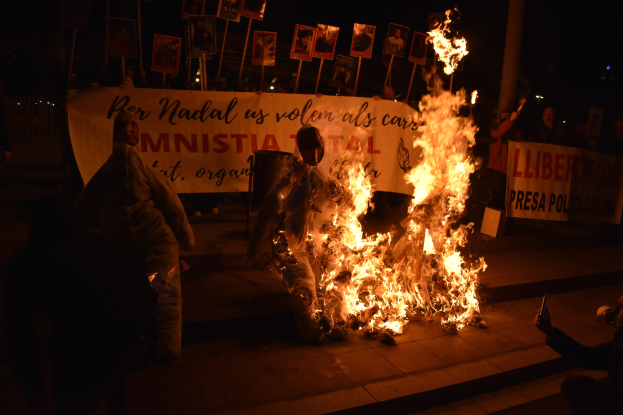 Gruppe von Menschen um ein hell loderndes Straßenfeuer versammelt, mit Schildern im Hintergrund und einem Müllcontainer im Vordergrund.