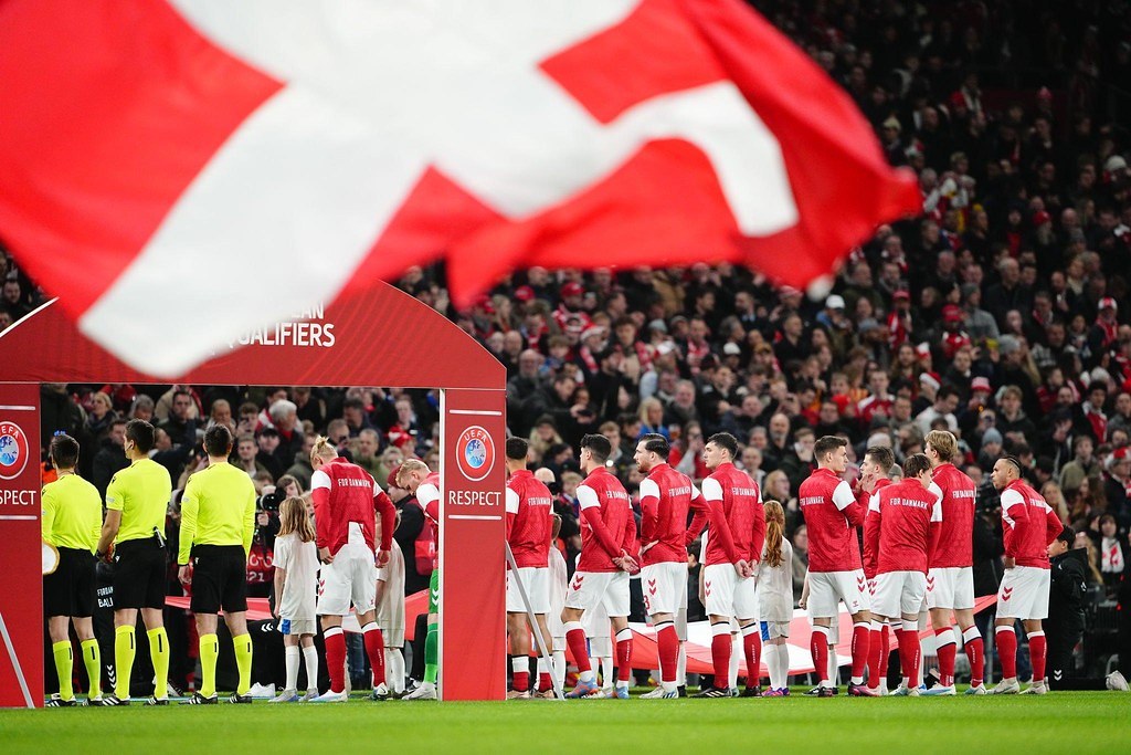 Gruppe von Menschen auf einem Fußballfeld mit einer roten und weißen Flagge im Vordergrund, einem Bogen mit "Bayern Munich vs Bayern Munich Betting Tips & Preview"-Text im Hintergrund und einer großen Menge im Stadion.