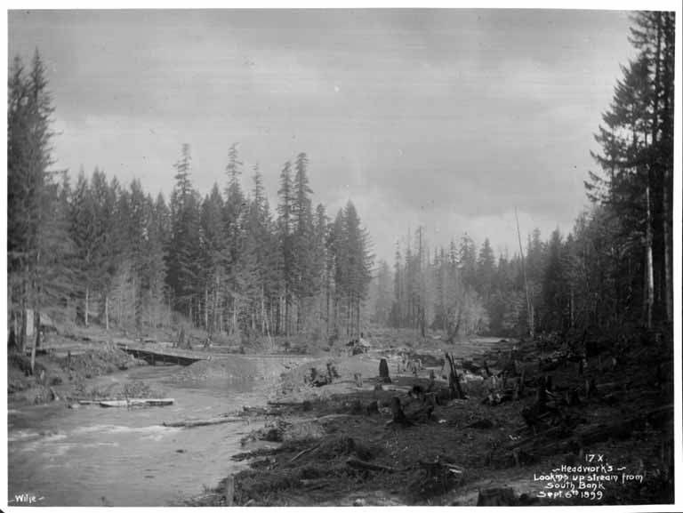 Ein Schwarz-Weiß-Foto eines Flusses im Wald, mit einem Kopfwerk, das von der Südseite stromaufwärts schaut, der Himmel im Hintergrund sichtbar und Text am unteren Bildrand.