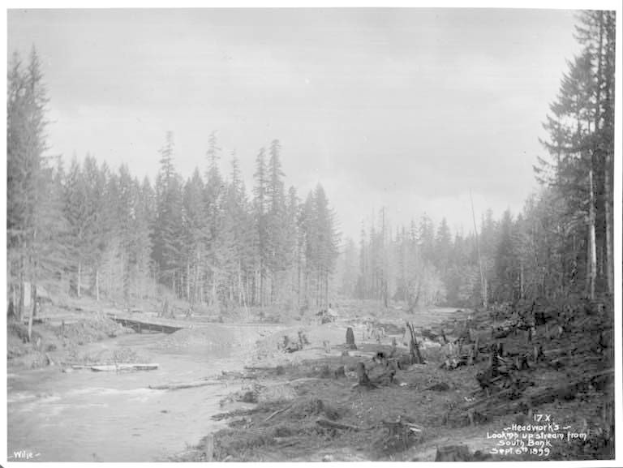 Ein Schwarz-Weiß-Foto eines Flusses im Wald, mit einem Kopfwerk, das von der Südseite stromaufwärts schaut, der Himmel im Hintergrund sichtbar und Text am unteren Bildrand.