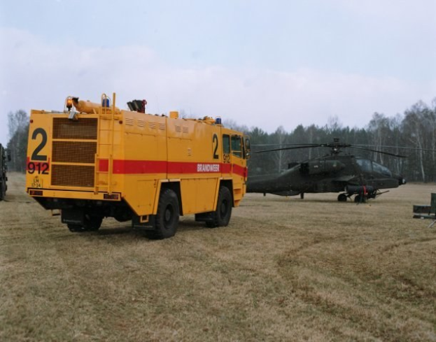 Ein gelbes Feuerwehrauto neben einem schwarzen Hubschrauber auf einer grünen Wiese geparkt, mit Bäumen im Hintergrund und einem klaren blauen Himmel darüber.