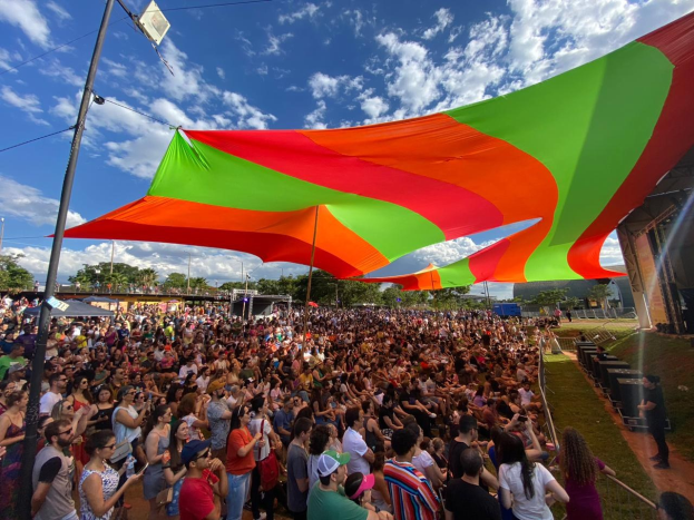 Eine große Menschenmenge steht vor einer Regenbogenflagge, mit einem Gebäude rechts daneben, Lautsprechern auf einer Bühne und Bäumen und Wolken am Himmel während einer Festival-Jubiläumsfeier.