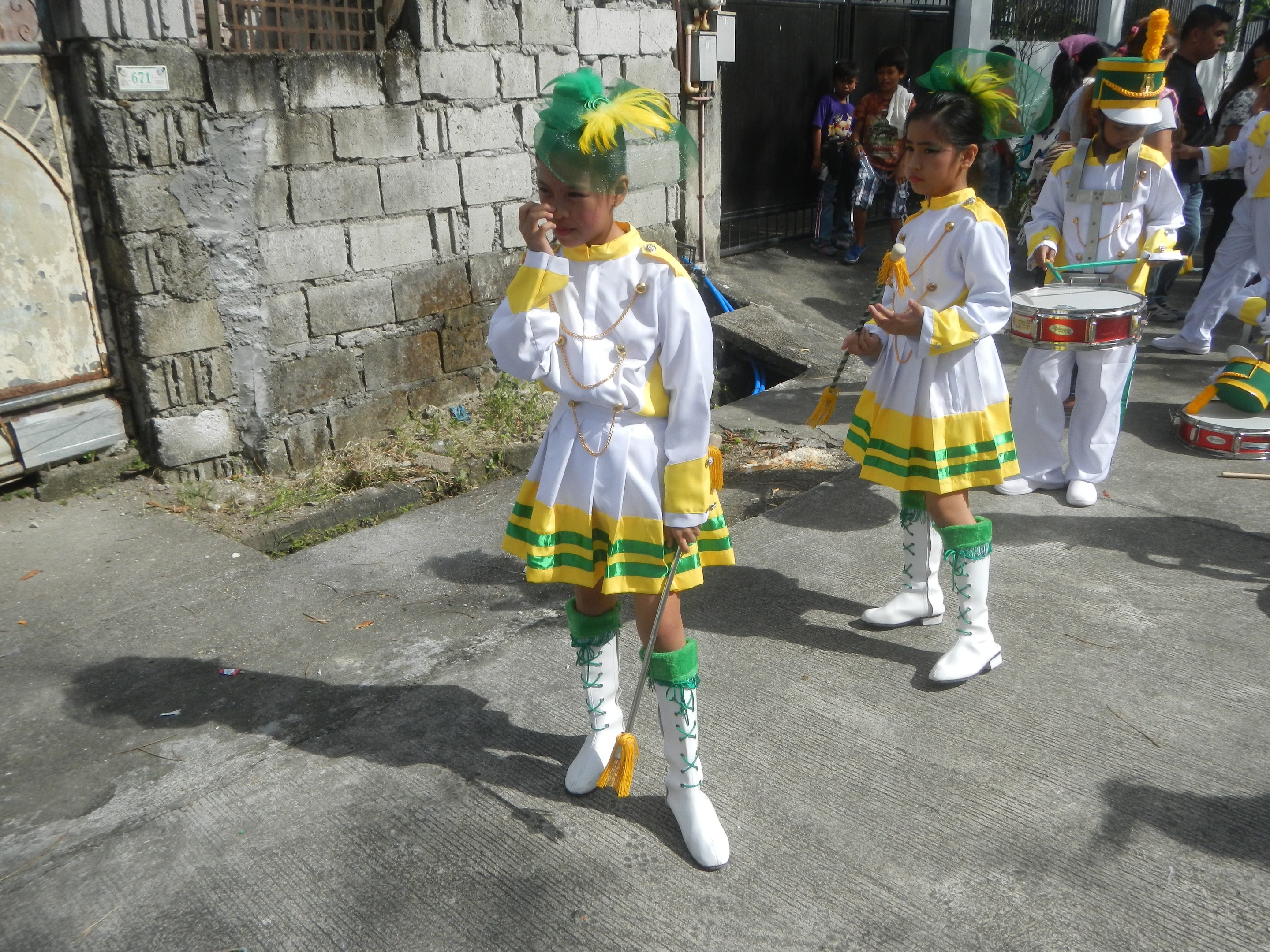 Gruppe junger Mädchen in weißen und gelben Outfits, die marschieren und Musikinstrumente spielen, vorbei an einer Wand, einem Tor und Bäumen.
